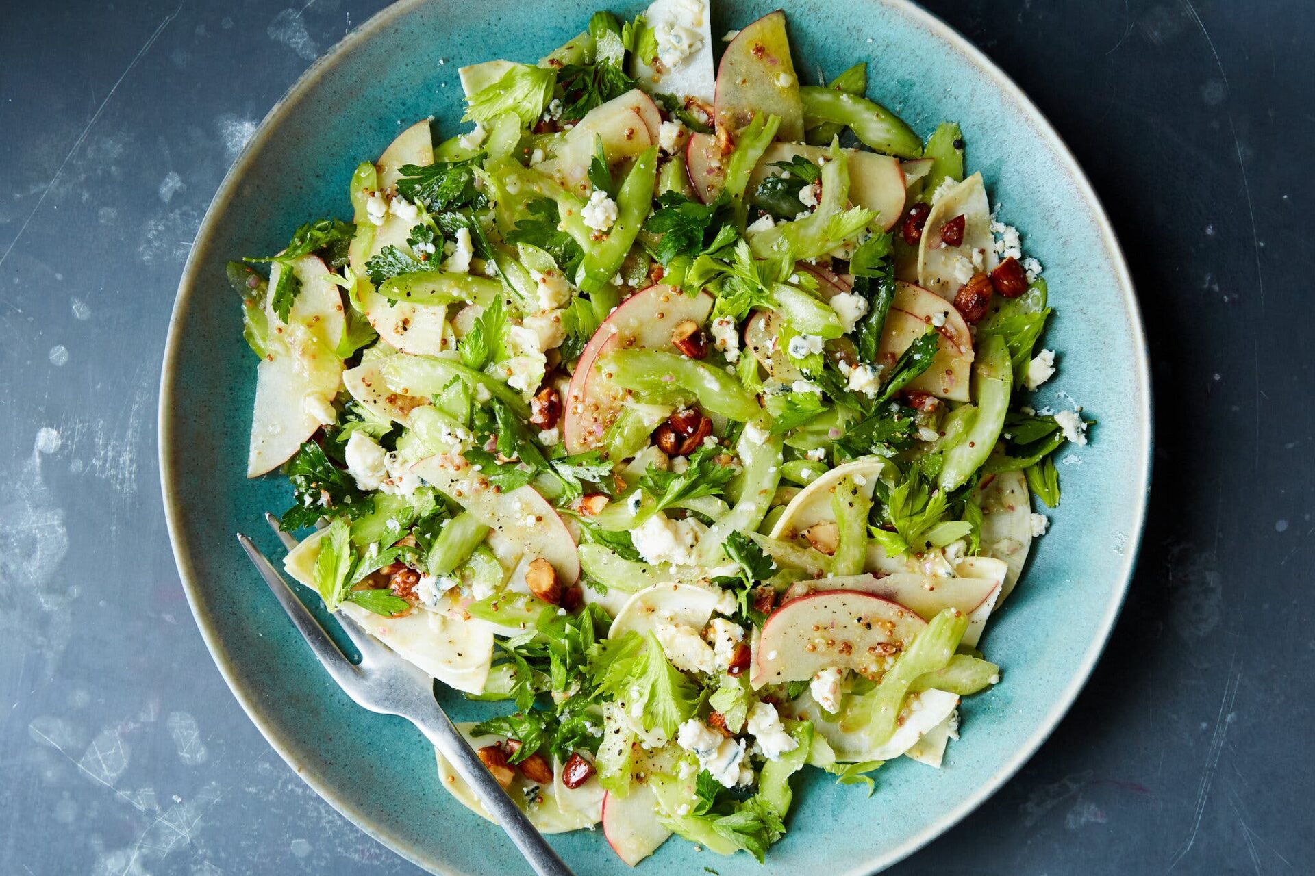 An overhead image of salad in a blue bowl, with sliced celery and celery leaves, sliced apples, chopped pecans and pieces of blue cheese. There is a fork in the bowl