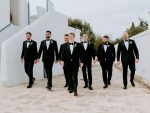 In questa foto, un gruppo di uomini in smoking nero con papillon cammina all’aperto in un contesto mediterraneo. La scena mostra diverse opzioni coordinate su come scegliere abito da sposo per il protagonista e i suoi testimoni.