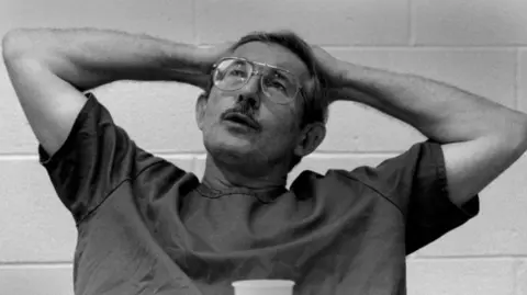 Getty Images Ames, in a black and white photo, puts both hands behind his head, with a white brick wall behind him